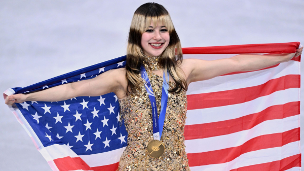 Alysa Liu wearing a gold skating costume wearing a gold medal and holding the American flag behind her